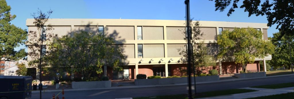 Wide view of a three-story concrete-and-brick building, its ground floor set back behind columns, partly screened by trees.