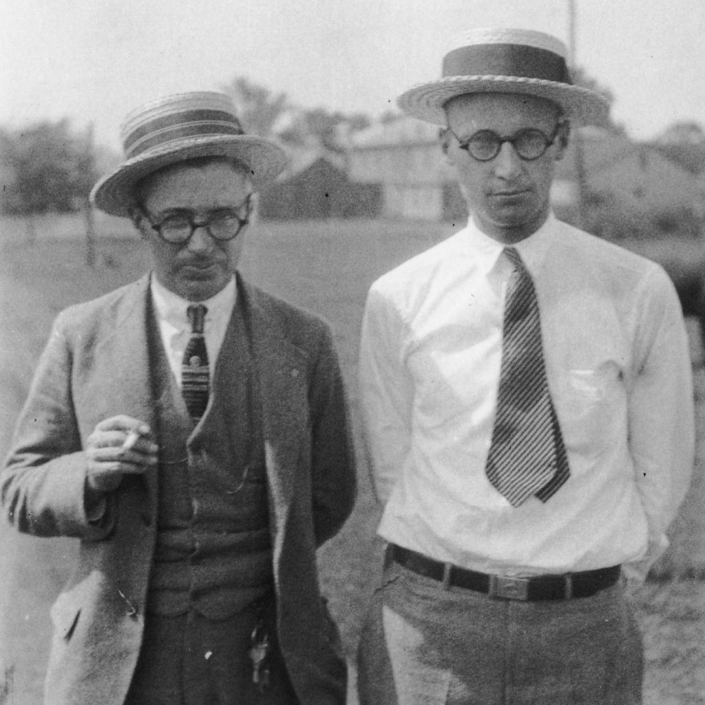 Two men in straw boater hats and round glasses standing outdoors. Rappleyea, left, wears a suit and holds a cigarette; Scopes, right, is in shirtsleeves and a striped tie.